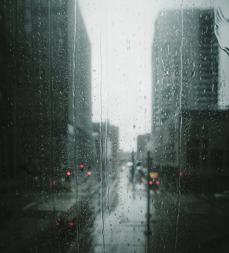 A view through a city window. The skies are grey amd the window coverd in rain, making it harder to see out. There are views of the city and traffic lights that are red.